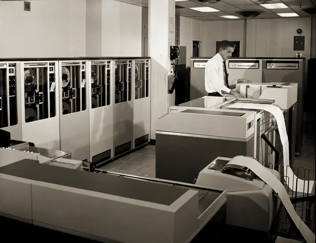 An operator at a line printer beside a bank of tape drives in a 1960s computer room